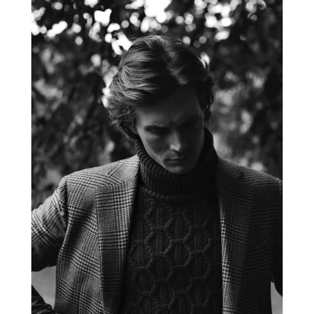 Monochrome portrait of a man with medium-length hair wearing a patterned blazer and a chunky knit turtleneck sweater outdoors