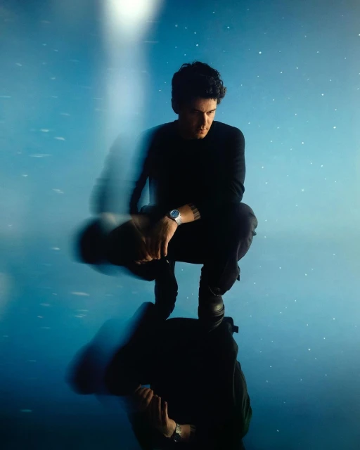 Portrait of a crouched man in black clothing with a watch, reflected in the water with a blue, starry sky and blurred light.