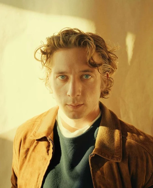 Close-up portrait of a fair-skinned adult male with wavy blonde hair, wearing a brown jacket and a blue sweater against a neutral background.