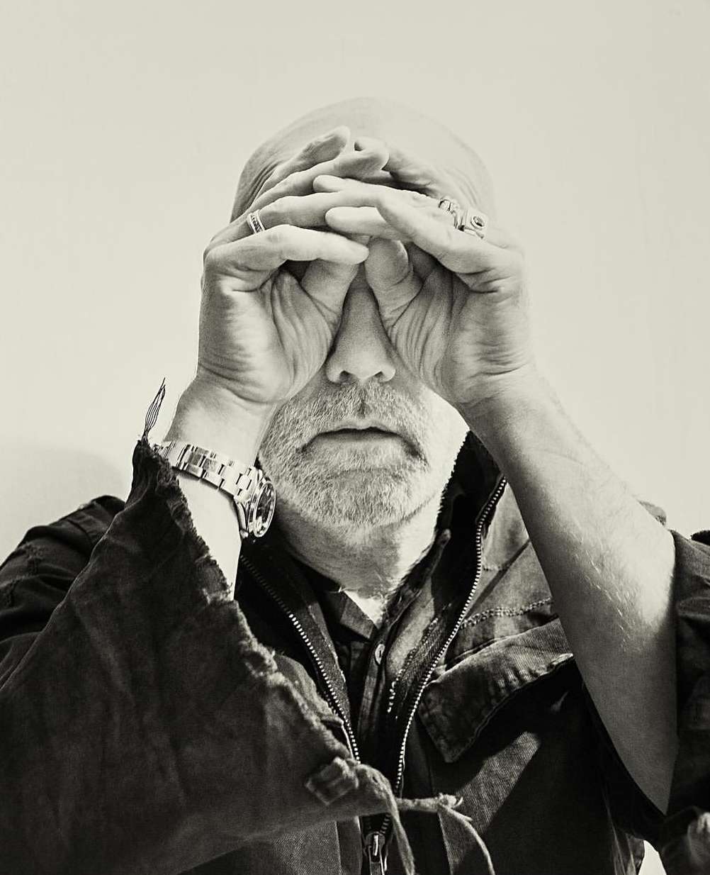 Close-up portrait of a bald man with a beard holding his hands to his face, wearing a dark jacket and a silver watch.