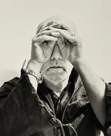 Close-up portrait of a bald man with a beard holding his hands to his face, wearing a dark jacket and a silver watch.