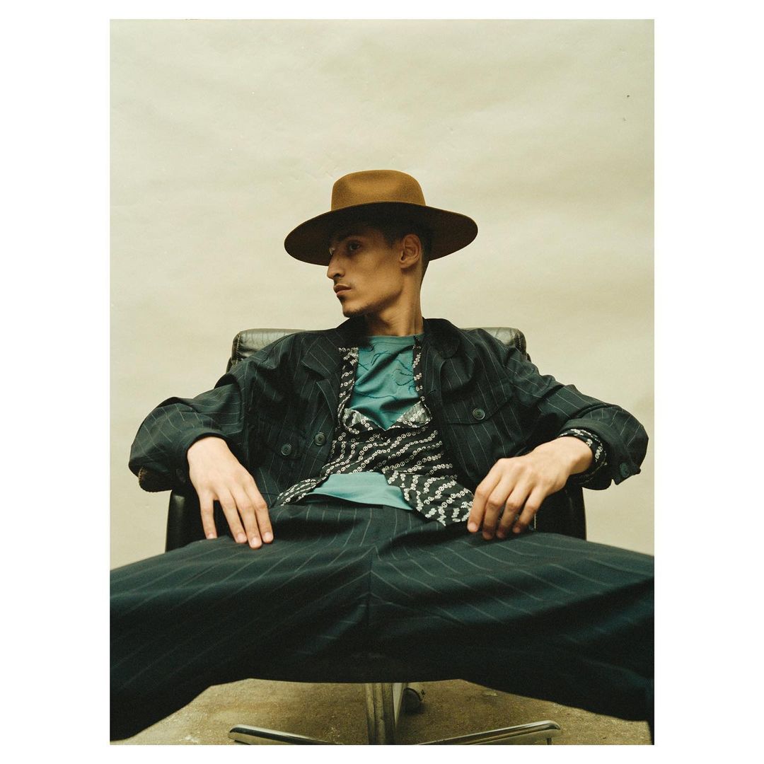 Portrait of a man in a pinstripe suit and brown hat, sitting in a black chair, looking left against a neutral studio background.