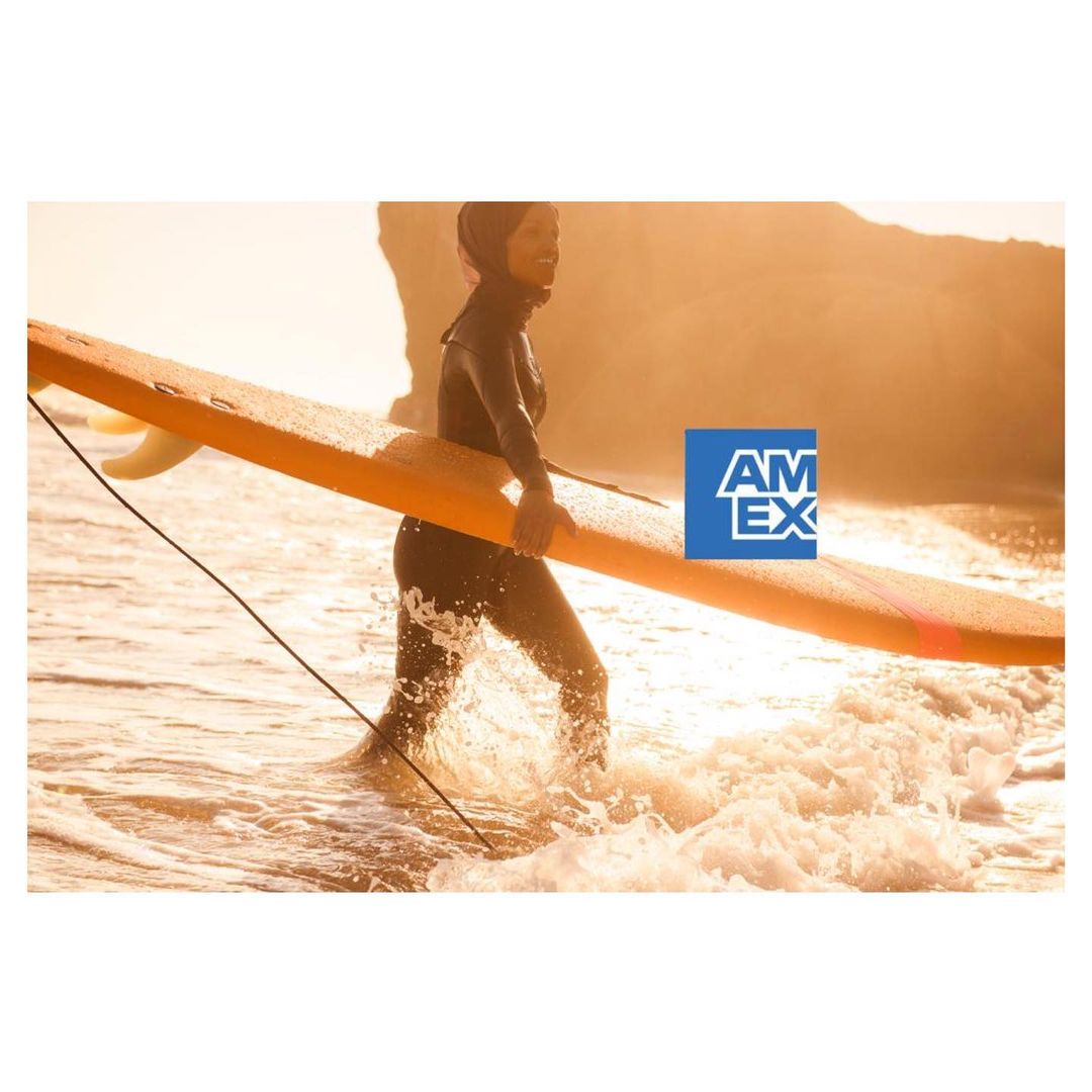 Woman in wetsuit carries orange surfboard in ocean with sandy beach and rock formations in background, with AMEX logo.