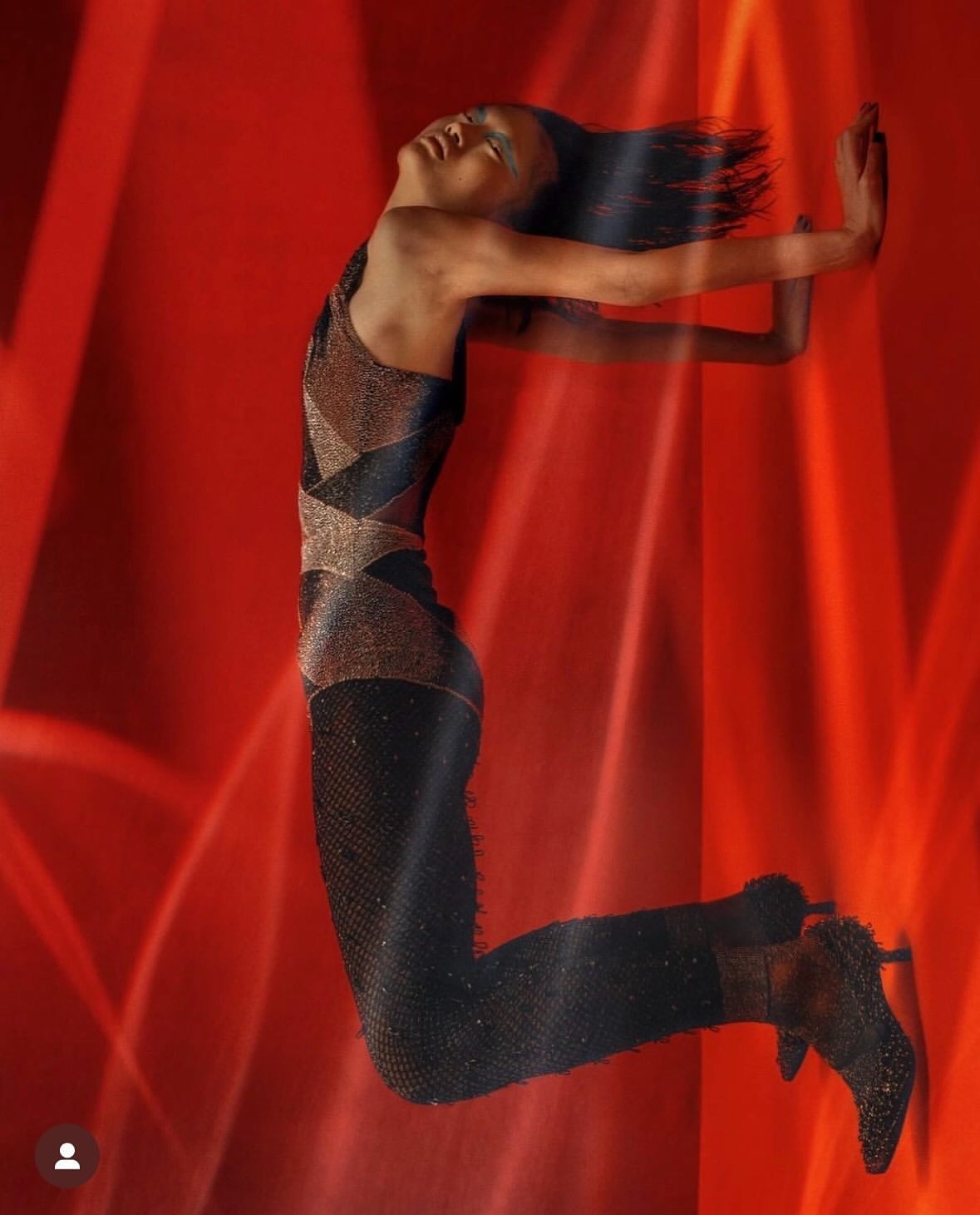 Fashionable woman with dark hair and sparkling jumpsuit in mid-air pose against red background with dynamic lighting effects for a high-fashion photo.