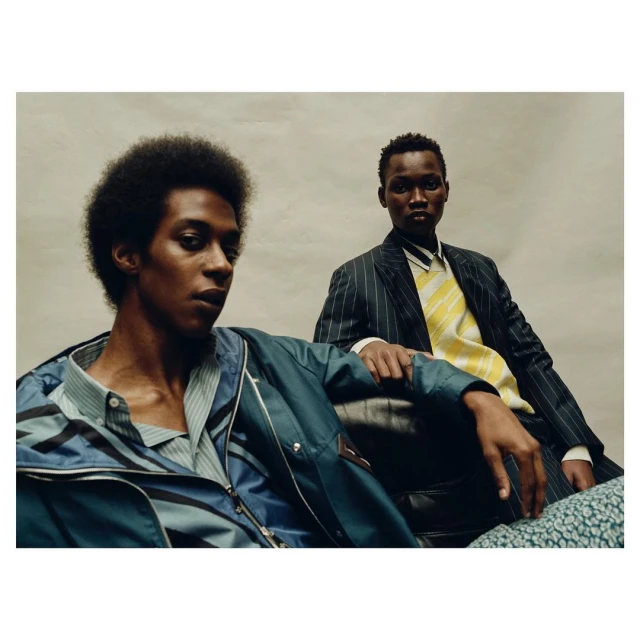 Two Black men pose indoors, modeling fashion jackets and blazers. One sits, one stands, in a studio setting with soft, diffused lighting.