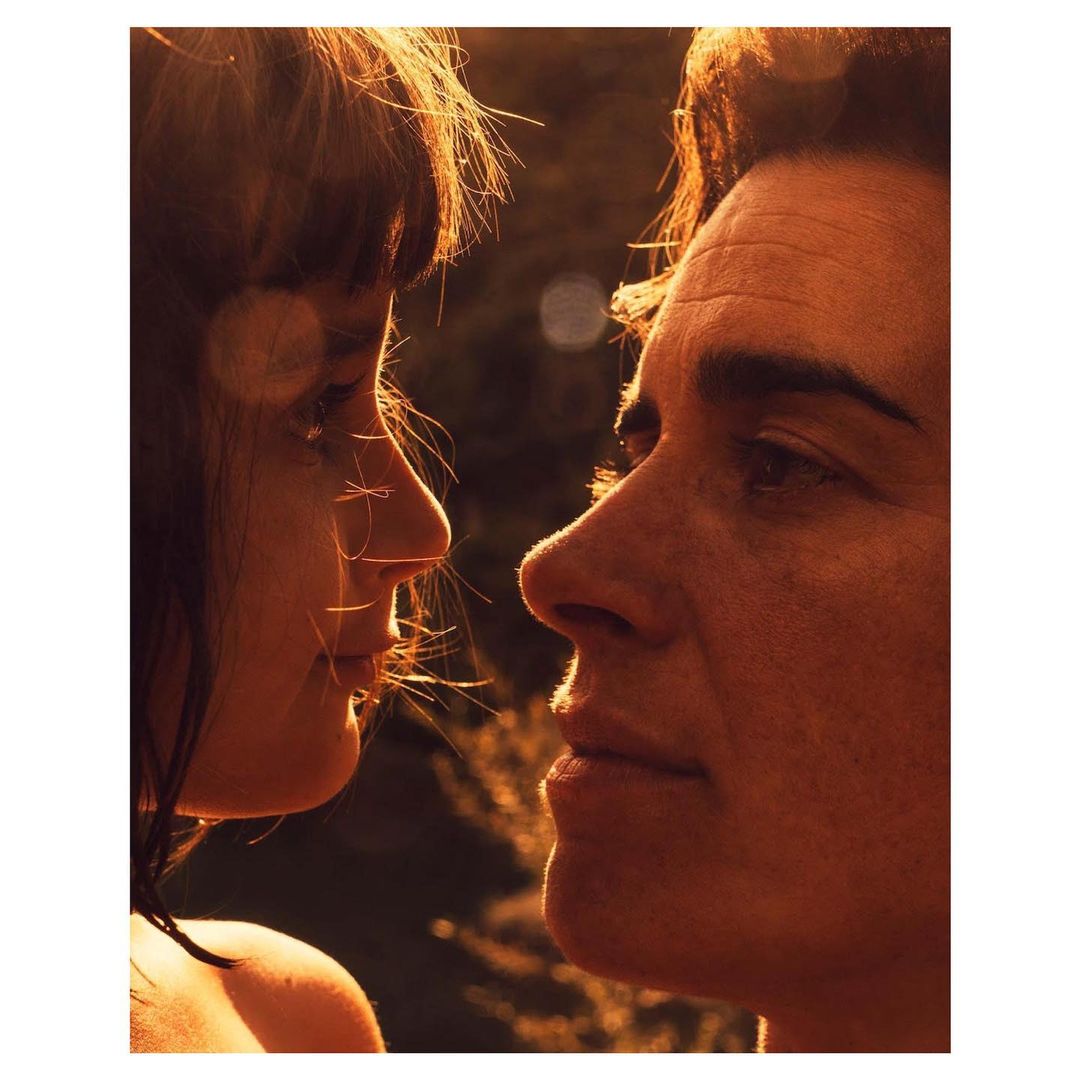 Close-up portrait of a couple, face to face, gazing at each other in warm, golden light with a soft, natural bokeh background of trees and foliage.