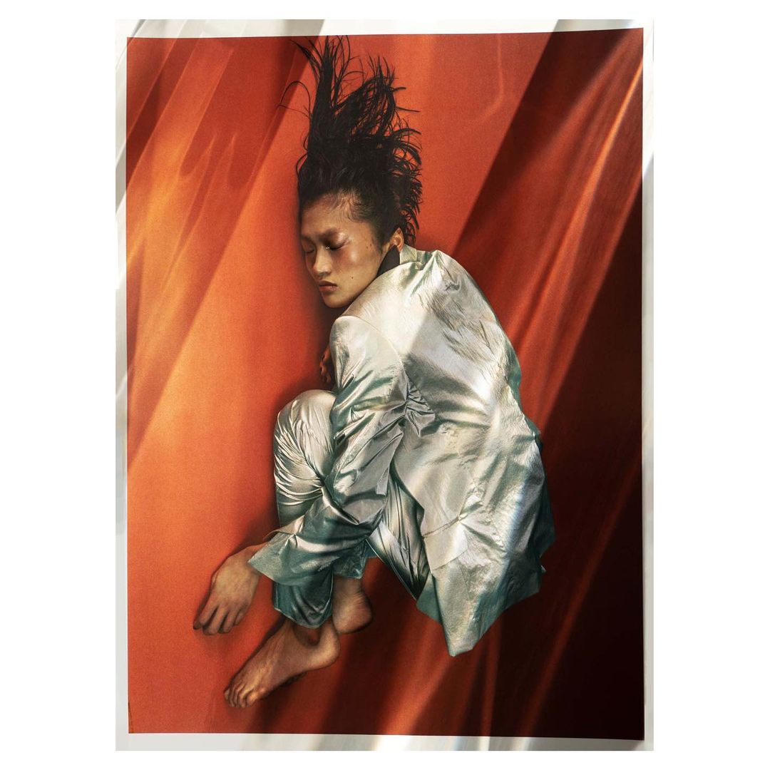 Woman in shiny suit poses against orange background with linear shadows. Fashion portrait features a unique hairstyle and bare feet.
