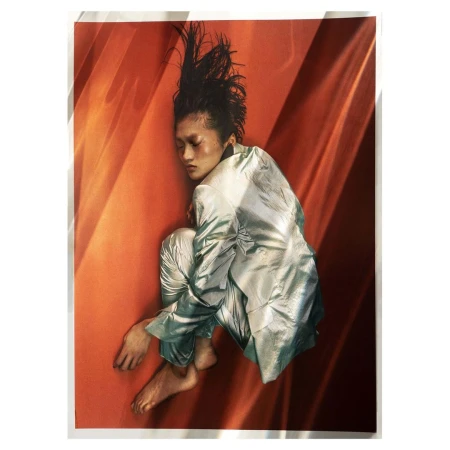 Woman in shiny suit poses against orange background with linear shadows. Fashion portrait features a unique hairstyle and bare feet.