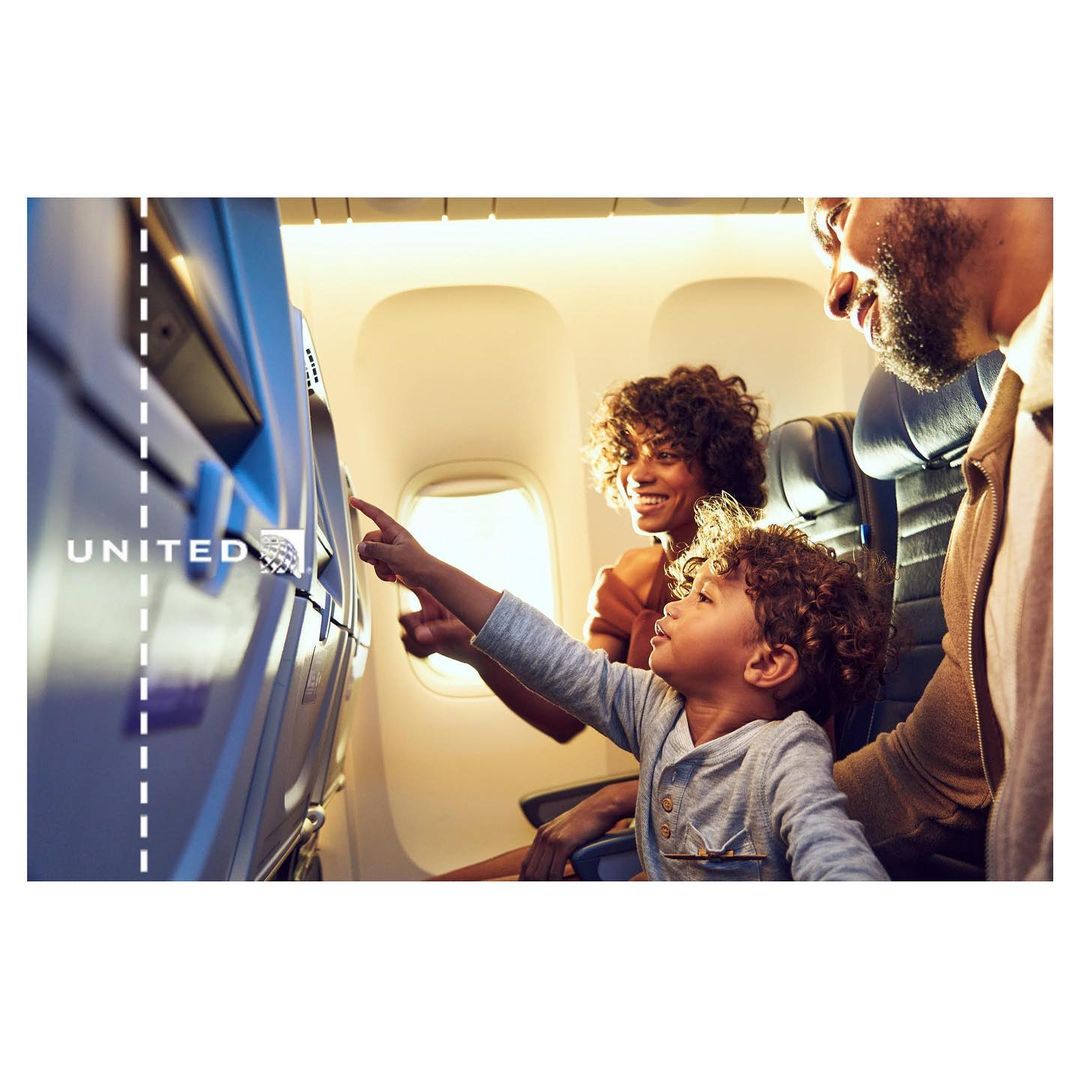 Family of three on an airplane, with a young child pointing at a screen, mother smiling, father looking on, representing air travel.