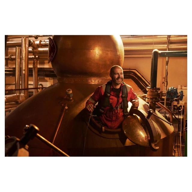 Man in red jumpsuit stands inside a copper vat in a distillery, surrounded by metal pipes and tanks, smiling in an industrial setting.