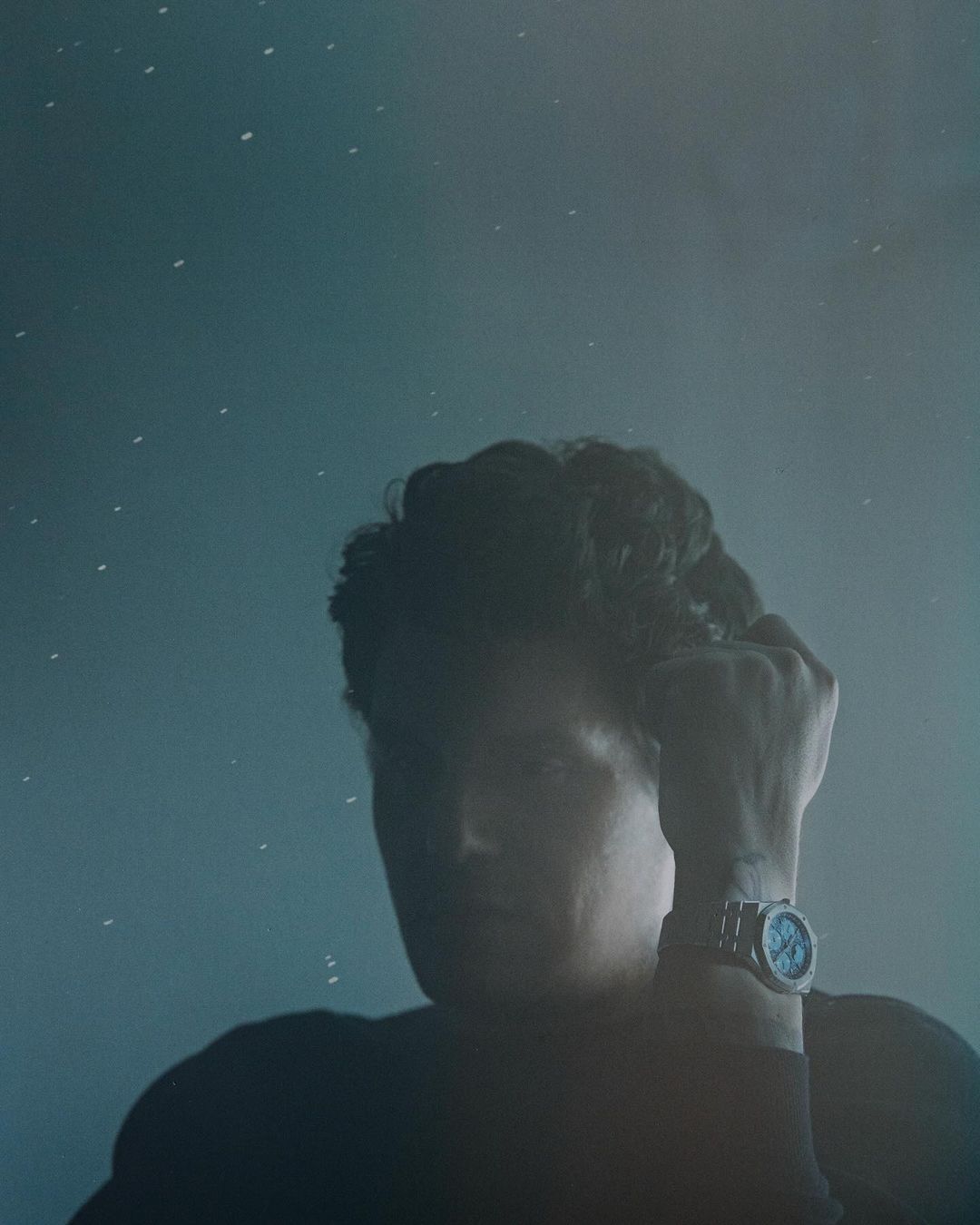Man with curly hair poses in a dark tee with a wristwatch, against a textured blue background speckled with tiny white dots.