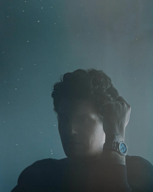 Man with curly hair poses in a dark tee with a wristwatch, against a textured blue background speckled with tiny white dots.
