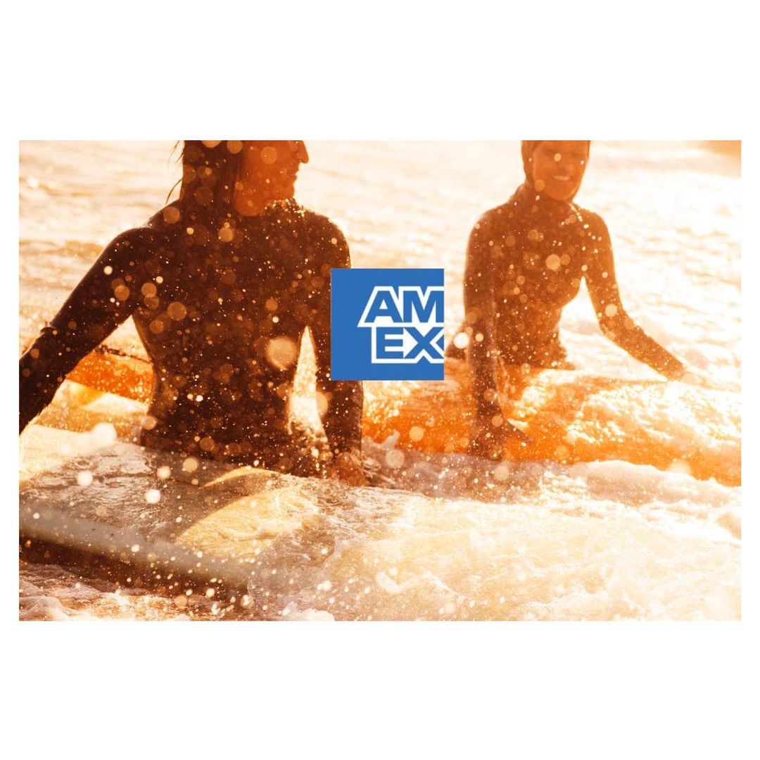 Two surfers on surfboards ride a wave toward the shore at sunset, with the American Express logo prominently displayed.
