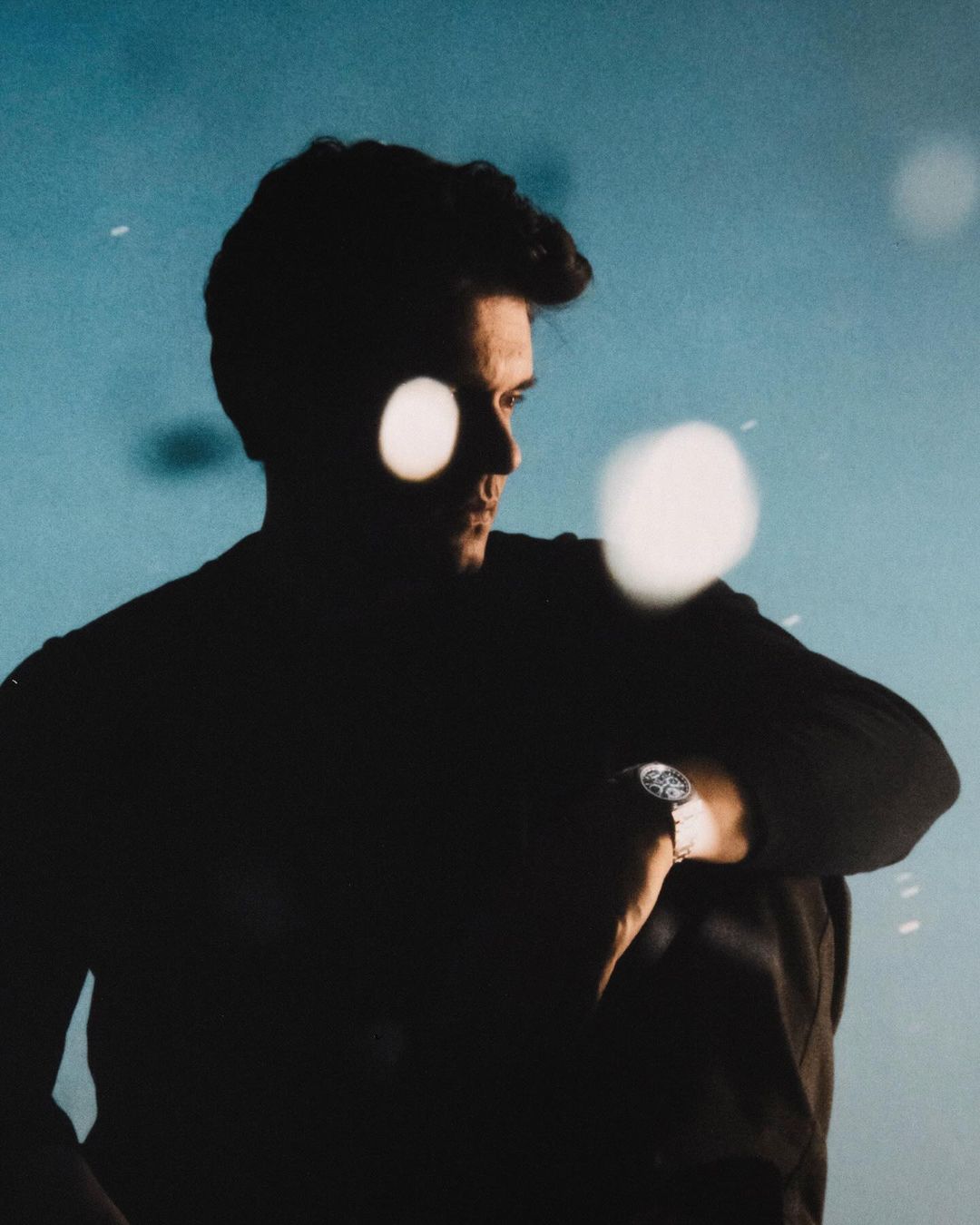 Dark-haired man checks wristwatch in a low-light setting with a blurry, light-blue background and bokeh circles adding a soft, hazy effect.