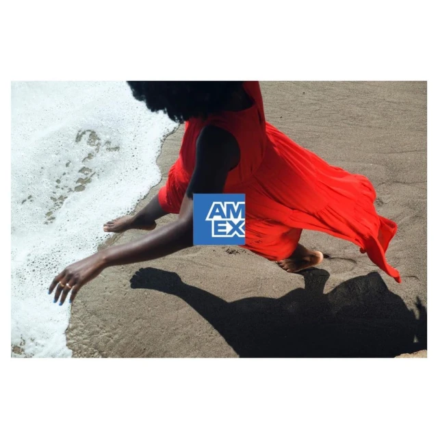 Woman in red dress sits on sandy beach near ocean waves. Shadow cast on sand. American Express logo visible on the side.