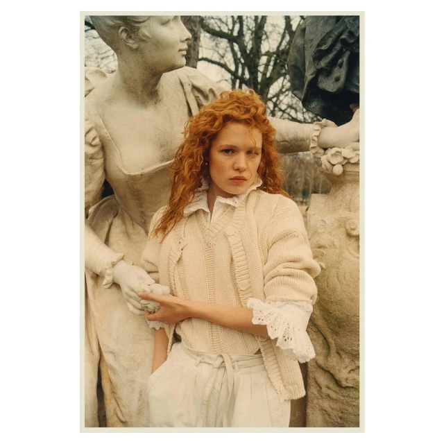 Red-haired woman in a cream cardigan stands next to a classical statue, outdoors in soft light, wearing white pants and a frilled collar.