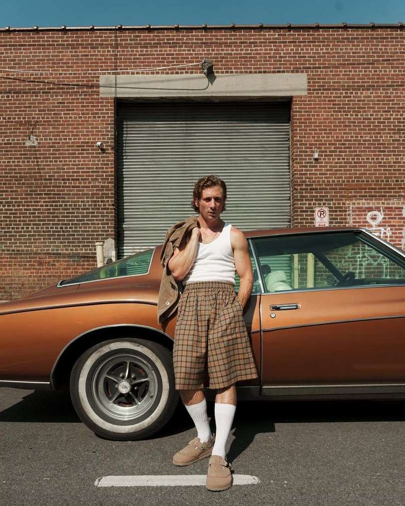 A man stands next to a vintage car in an urban setting, wearing a white tank top, plaid shorts, and tan loafers with a jacket over his shoulder.