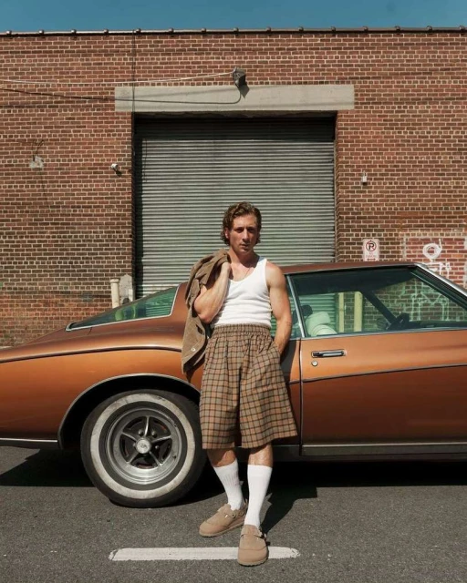 A man stands next to a vintage car in an urban setting, wearing a white tank top, plaid shorts, and tan loafers with a jacket over his shoulder.
