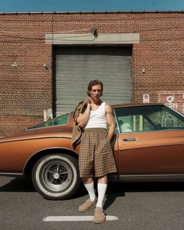 A man stands next to a vintage car in an urban setting, wearing a white tank top, plaid shorts, and tan loafers with a jacket over his shoulder.