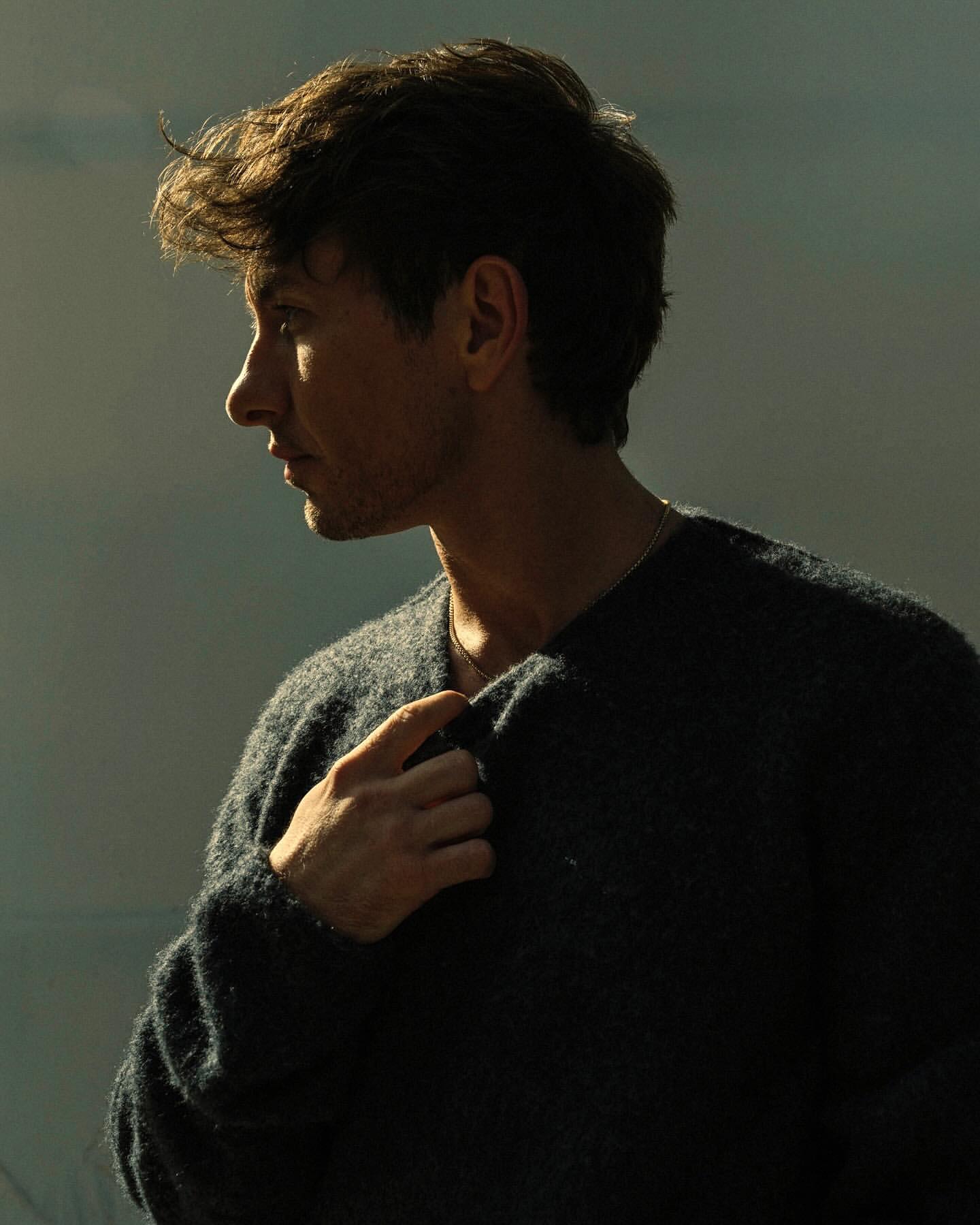 Portrait of a handsome man in a dark sweater, side profile with blurred background, warm lighting, and shallow depth of field.