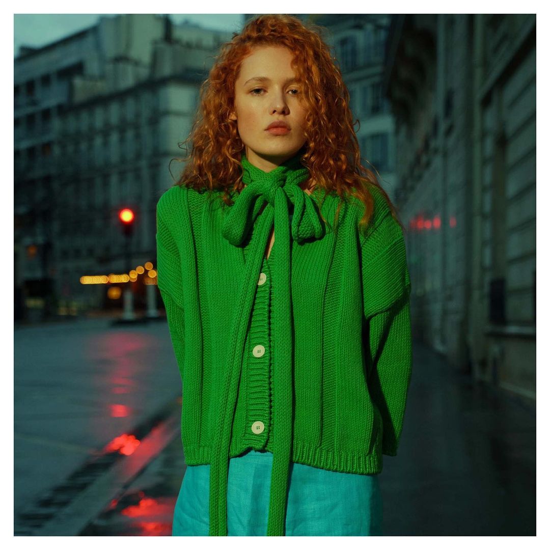 Adult woman with curly red hair poses in a green knit cardigan and blue pants in front of buildings on a city street at night.