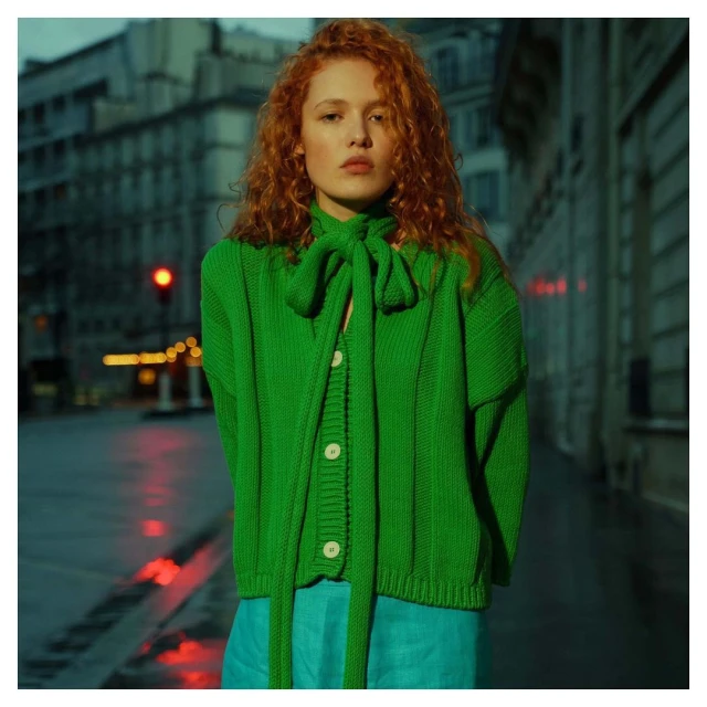 Adult woman with curly red hair poses in a green knit cardigan and blue pants in front of buildings on a city street at night.
