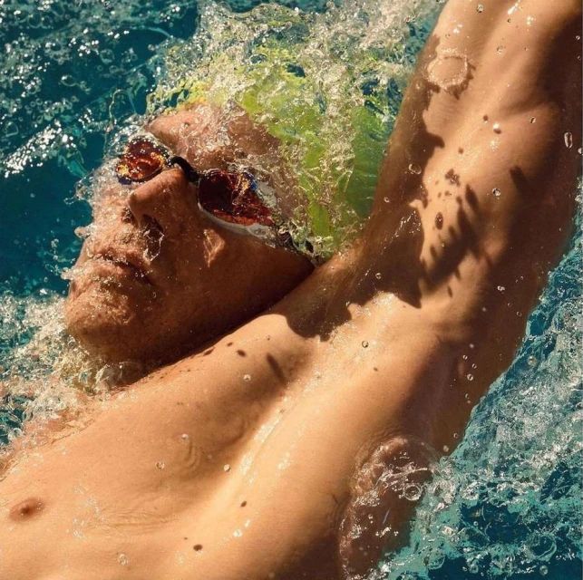 Close-up of a swimmer with a green cap, goggles, and shoulder out of the water in a blue-tiled pool, showcasing fitness and sport.
