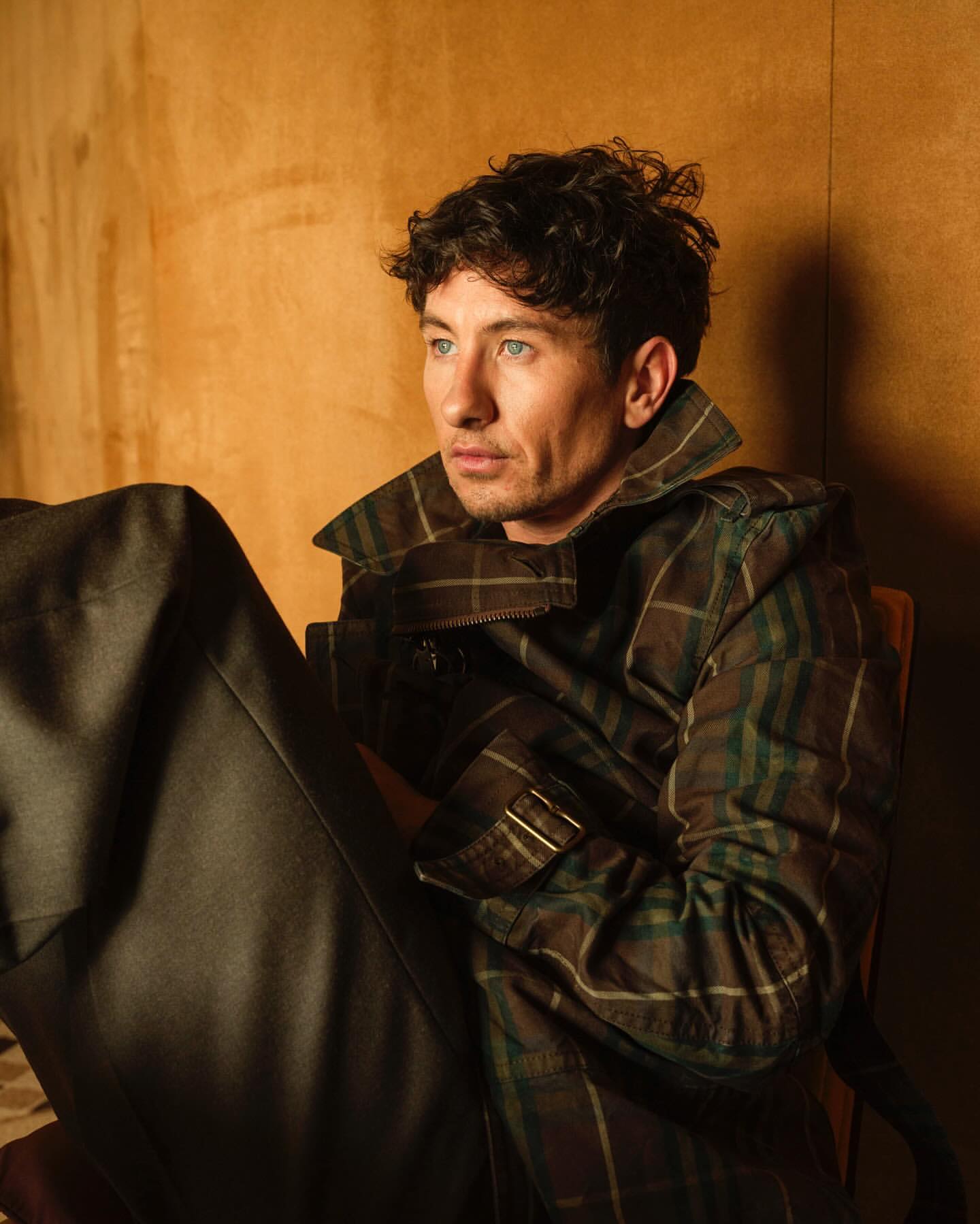 A man with curly dark hair sits in a chair looking off camera, wearing a plaid jacket and dark pants in a soft lit indoor setting.