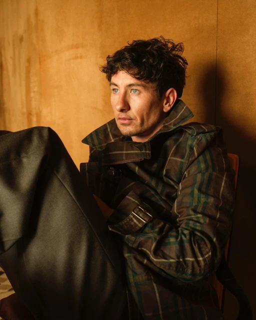 A man with curly dark hair sits in a chair looking off camera, wearing a plaid jacket and dark pants in a soft lit indoor setting.