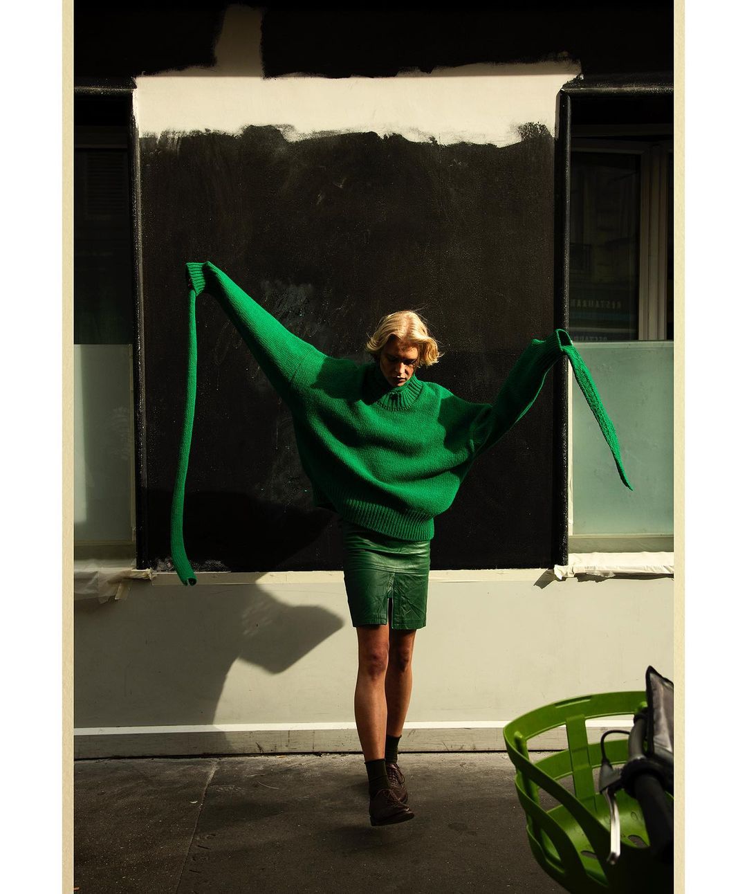 Fashion photo of a blonde adult wearing a green sweater and skirt standing outdoors with arms outspread against a geometric background.
