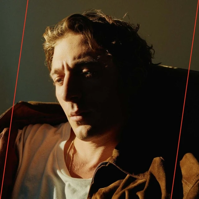 Close-up of a young adult male with wavy brown hair, wearing a white t-shirt and brown suede jacket, partially illuminated by natural sunlight.