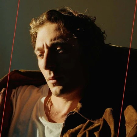 Close-up of a young adult male with wavy brown hair, wearing a white t-shirt and brown suede jacket, partially illuminated by natural sunlight.