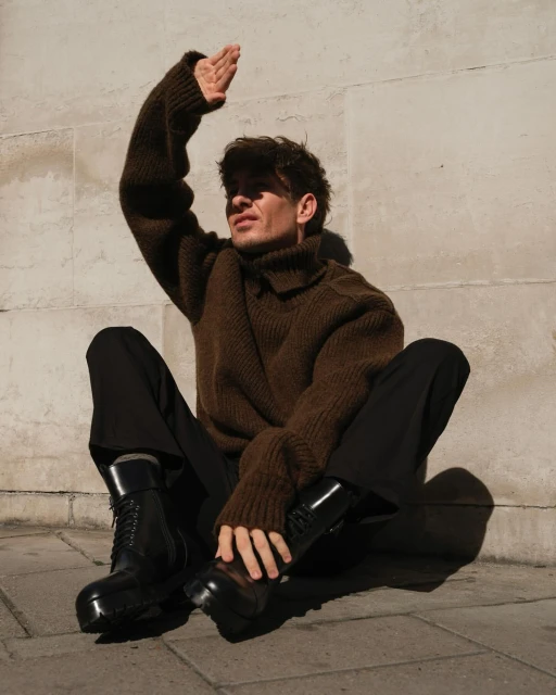 A young adult male sits outside on stone with his hand up, blocking the sunlight. He is wearing a brown sweater and black pants and boots.
