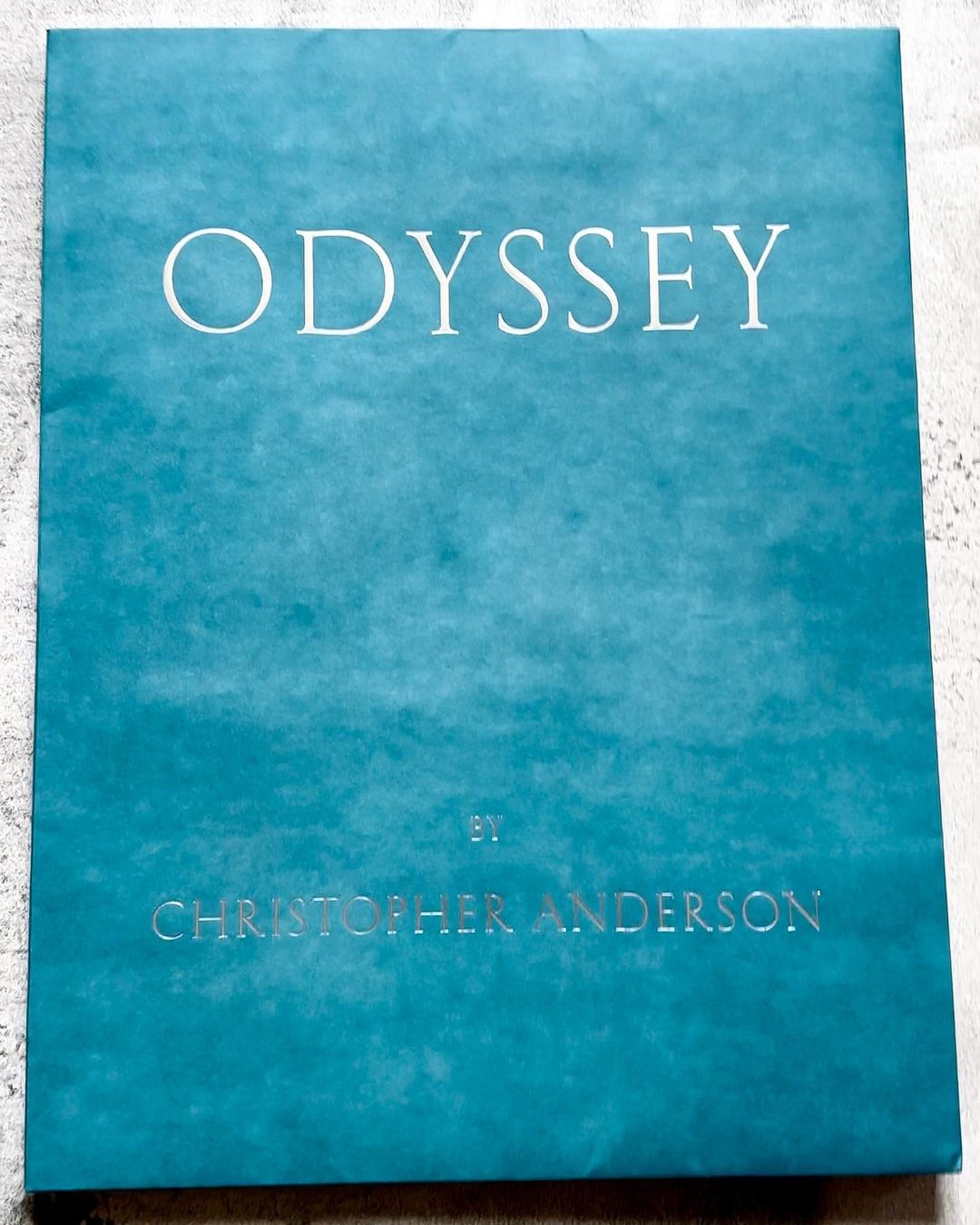 Close-up studio shot of a turquoise book cover with the word 'ODYSSEY' and author's name 'CHRISTOPHER ANDERSON' printed in white.