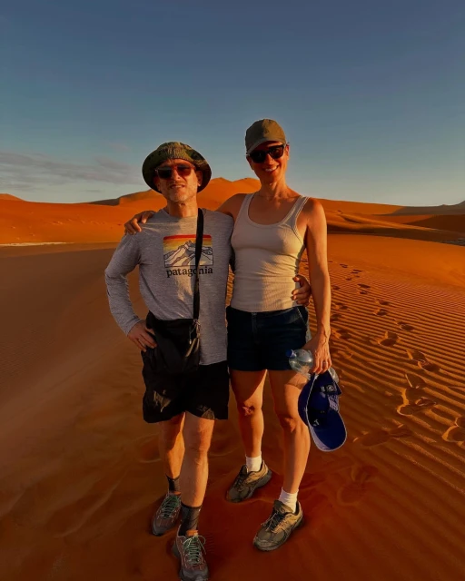 Two adults pose on a sand dune with a backdrop of desert hills under a clear sky, creating a portrait in a natural setting.
