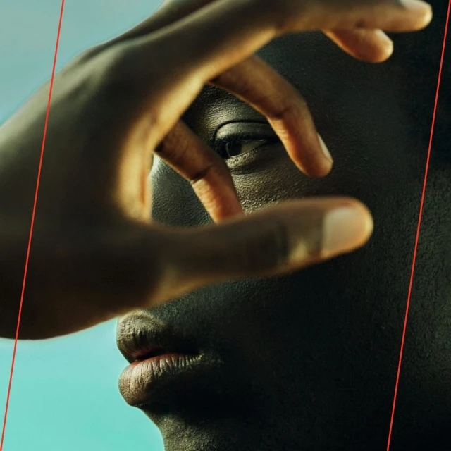 Close-up of an adult's dark-skinned face with their hand framing their eye, set against a light blue background with visible studio lighting.