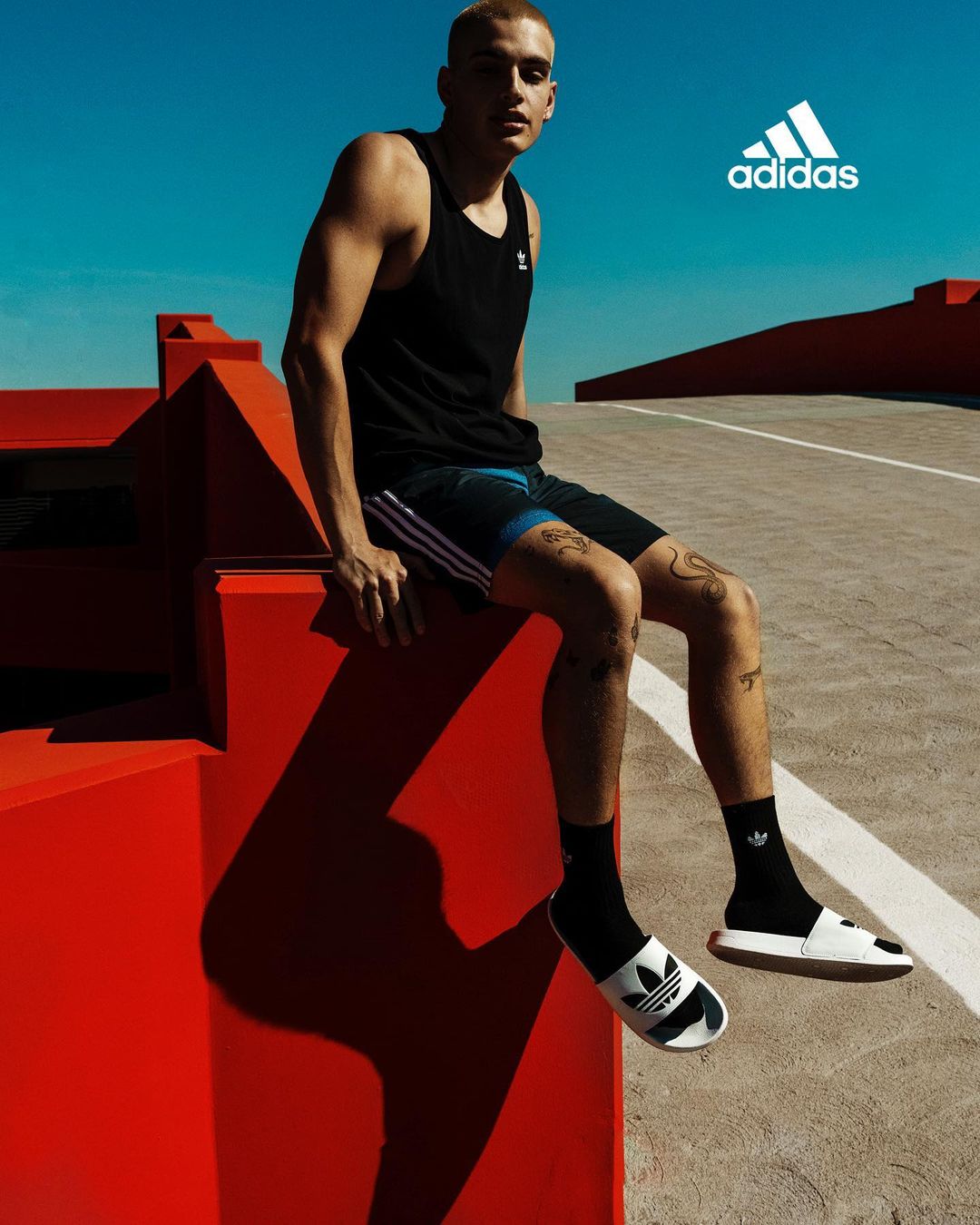 Man in Adidas activewear sits on an orange structure against a blue sky, showcasing a sporty and relaxed outdoor lifestyle.