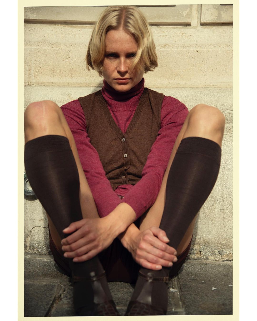 Blonde woman poses seated outdoors wearing brown vest, plum turtleneck, knee-high socks, and Mary Jane shoes in soft sunlight.