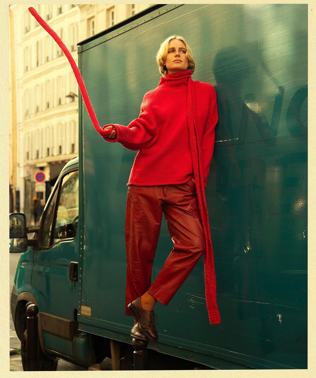 Fashion shot of a blonde woman in a red sweater and leather pants standing next to a dark green van in an urban environment.