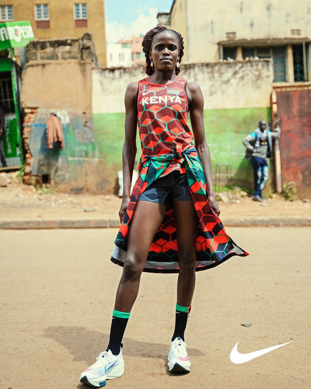 Female athlete in Kenya racing uniform stands on a dirt road in front of buildings, wearing running shoes with the Nike logo visible.