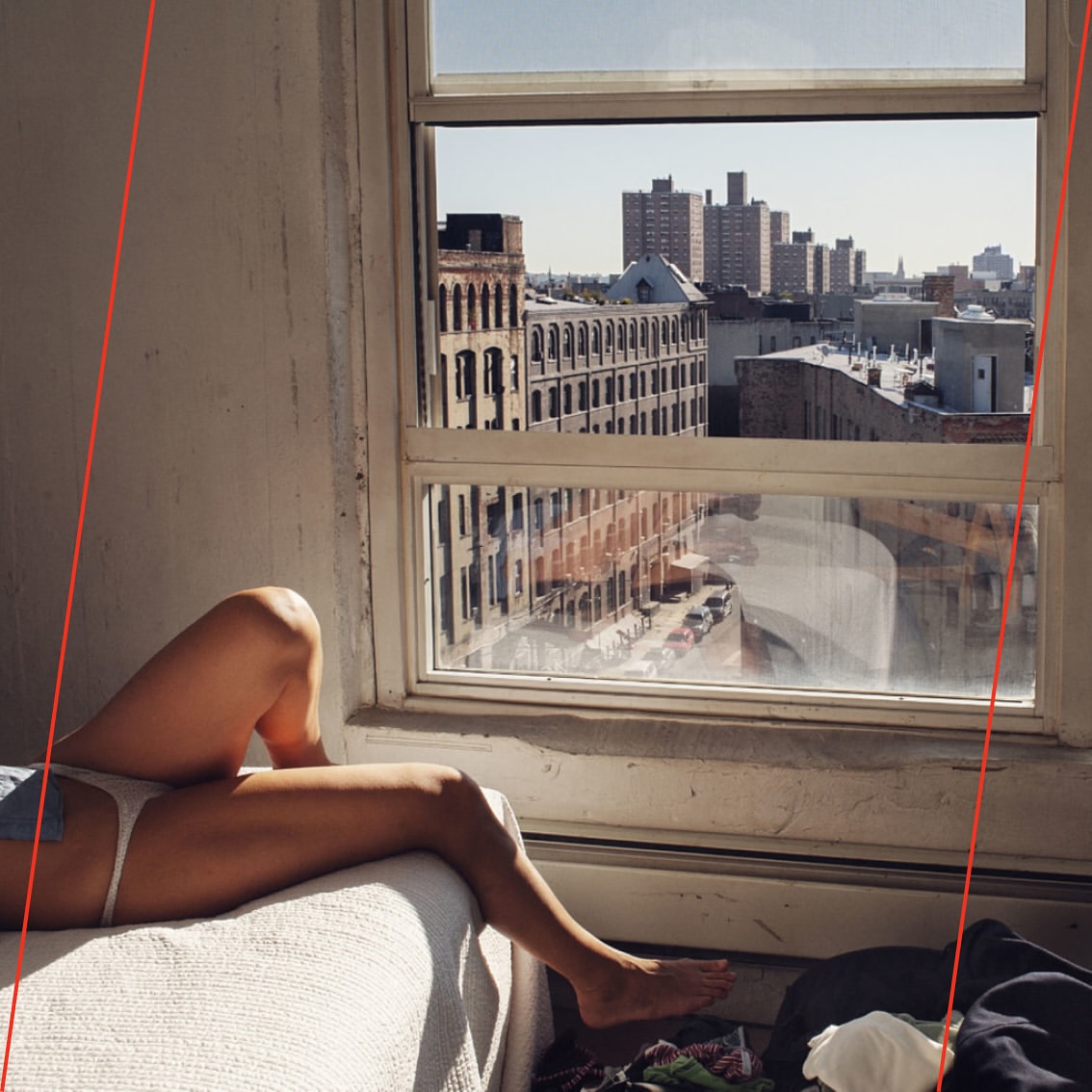 Woman lounges by a window with an expansive cityscape view, bathed in natural light. Interior shows a casual, lived-in space.
