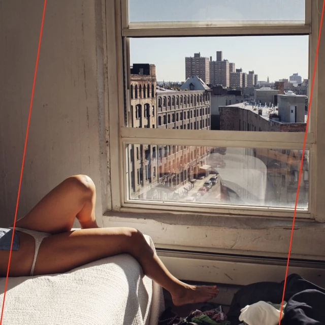 Woman lounges by a window with an expansive cityscape view, bathed in natural light. Interior shows a casual, lived-in space.
