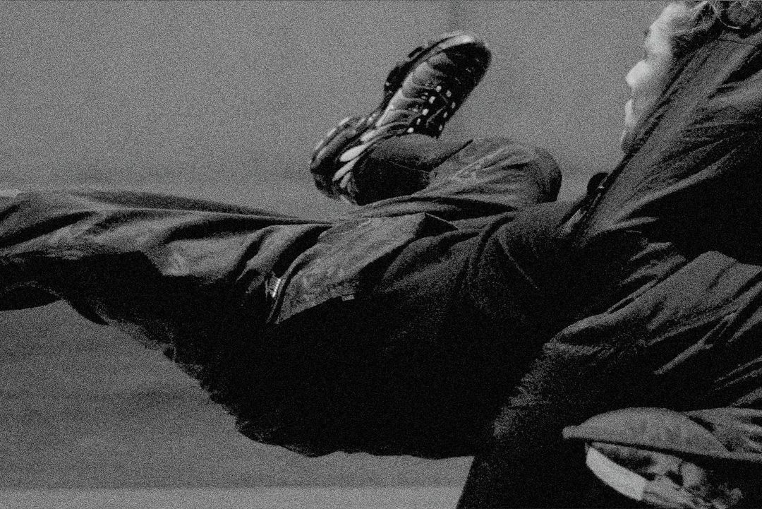 Black and white image of a person wearing a coat and pants, with one leg raised in the air showing the sole of their shoe.