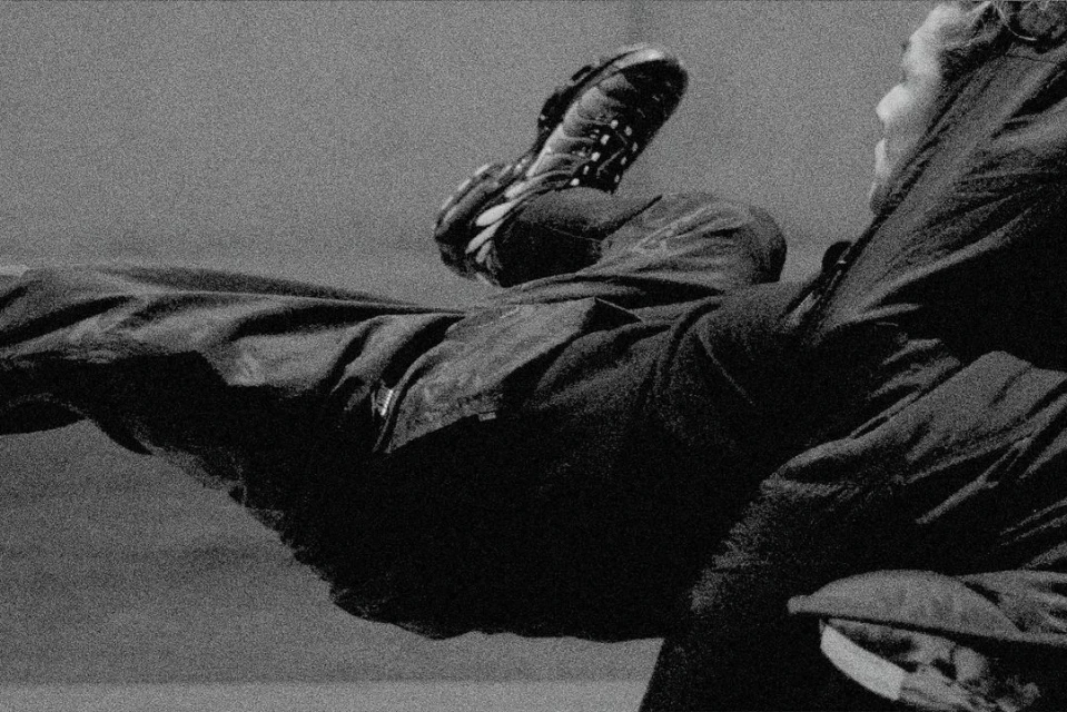 Black and white image of a person wearing a coat and pants, with one leg raised in the air showing the sole of their shoe.