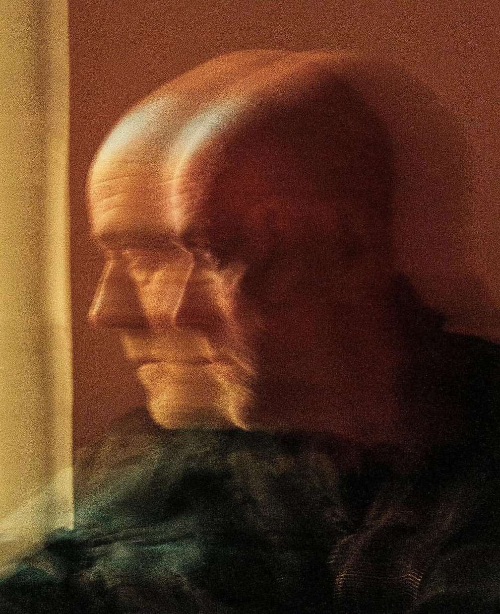 Blurred image of a bald adult man in profile, wearing a dark top, with a yellow wall in the background. Motion blur is evident.