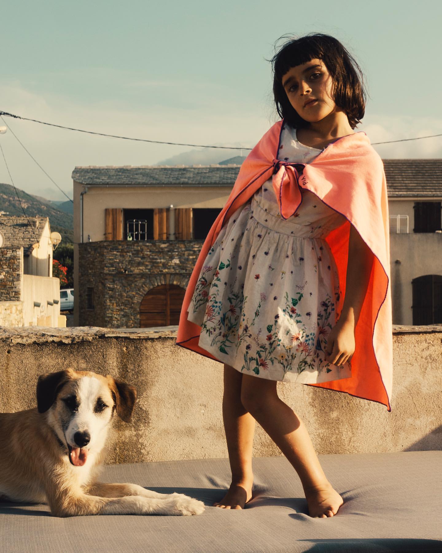 Girl in floral dress and coral cape poses outdoors with dog, set against a backdrop of houses and mountains on a sunny day.