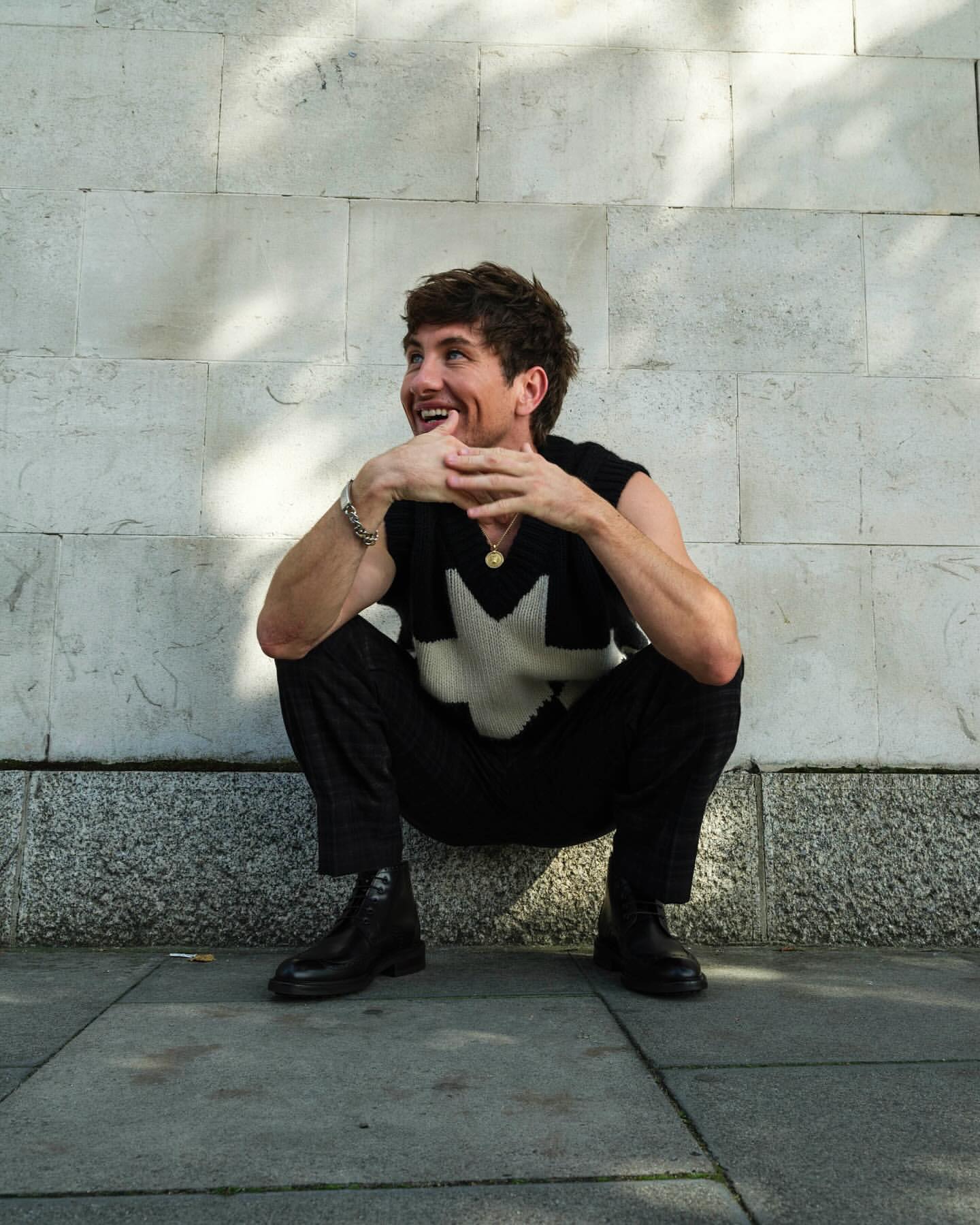 A smiling adult man in a sleeveless top squats outdoors against a textured wall, hands clasped, wearing black pants and black leather boots.