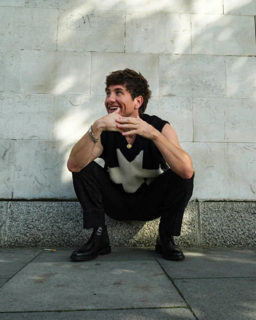 A smiling adult man in a sleeveless top squats outdoors against a textured wall, hands clasped, wearing black pants and black leather boots.