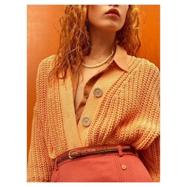 Close-up of an adult female wearing an orange knit cardigan, pearl necklace, and belted skirt against a solid orange background.