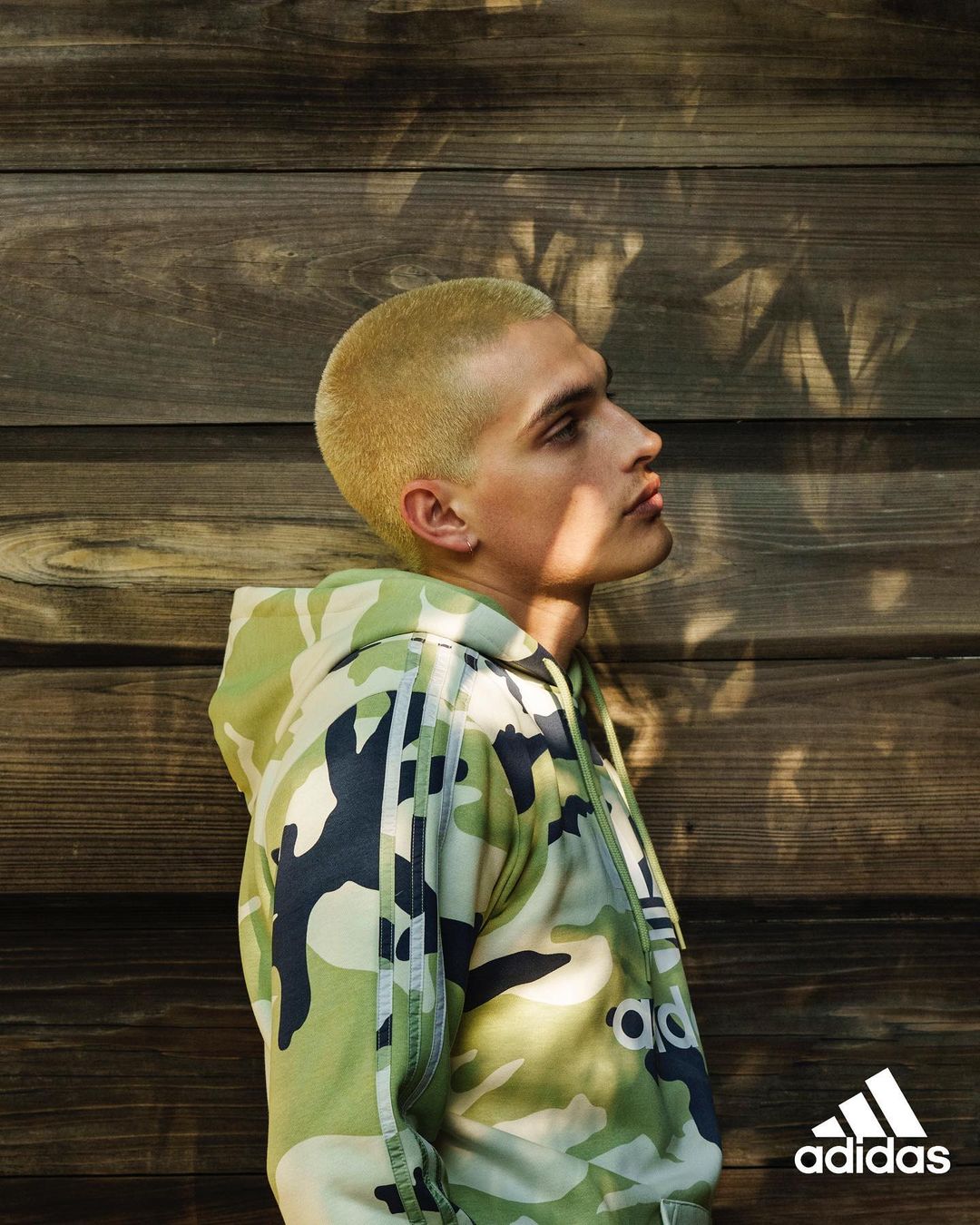 Profile shot of a young adult wearing a camo-patterned Adidas hoodie against a wood panel wall, showing a fashion or lifestyle scene.